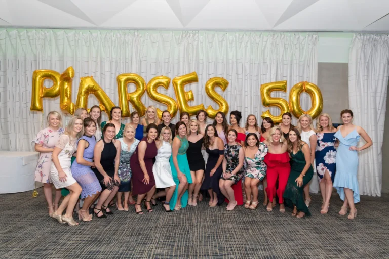 The image captures a large group of women posing together at the Pearses 50th Anniversary Awards Night. Some are standing in the back while others kneel in front, all smiling at the camera. They wear a variety of formal dresses in different colors, styles, and patterns. Behind them, a large gold balloon sign reads "PEARSES 50," highlighting the anniversary celebration. The indoor venue features a pleated light-colored curtain backdrop and a patterned carpet. The atmosphere is festive, joyful, and social, reflecting a celebratory group moment.