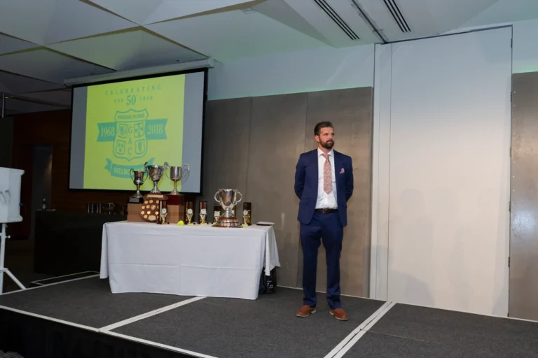 This photograph, taken during the Pearses 50th Anniversary Awards Night at Zinc, Fed Square, shows a man standing on stage in a blue suit with a patterned tie and pocket square, hands clasped behind his back, looking thoughtfully to the side. To his left, a long table displays trophies and awards, including large silver cups and smaller plaques. The background features a gray paneled wall and a projection screen displaying the event logo, "CELEBRATING OUR 50 YEARS 1968-2018 PEARSES MELBOURNE." The atmosphere is formal and poised, capturing a reflective moment during the awards presentation.