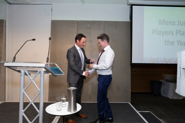 This photograph, taken at the Pearses 50th Anniversary Awards Night at Zinc, Fed Square, shows two men on stage shaking hands as an award is presented. The man on the left, wearing a dark gray suit and striped tie, hands a medal or small trophy to the man on the right, dressed in a white shirt, dark tie, and blue trousers, who smiles at the presenter. Behind them, a large screen displays partial text for the "Mens Junior Players Player of the Year" award. A clear podium and a small table with a pitcher and glasses are visible, highlighting the formal and celebratory atmosphere of the awards presentation.