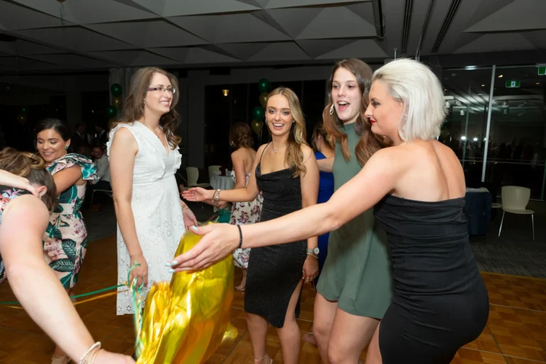 A lively moment at the Pearses 50th Anniversary Awards Night shows a group of women dancing and laughing on a dark wood dance floor. One woman in a white lace dress holds a large gold balloon shaped like a number, while others in black, green, and sparkly dresses smile and reach toward it. The modern event space features round tables with white tablecloths, clusters of green and gold balloons, and warm lighting. The atmosphere is energetic, joyful, and celebratory, capturing the fun and friendship of the occasion.