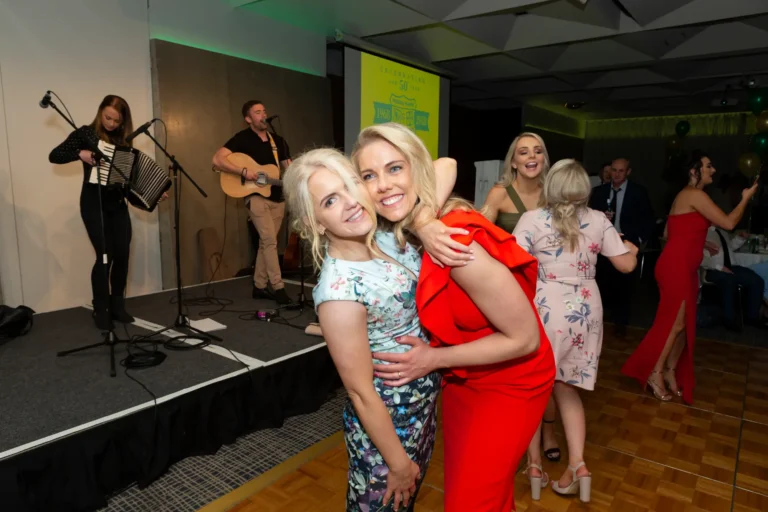 A candid and joyful moment at the Pearses 50th Anniversary Awards Night, showing two women embracing and smiling at the camera. One wears a red ruffled dress, the other a light floral dress. Guests dance and a live band plays on stage in the background, with gold and green balloons adding a festive touch. The atmosphere is lively, celebratory, and full of friendship and happiness.