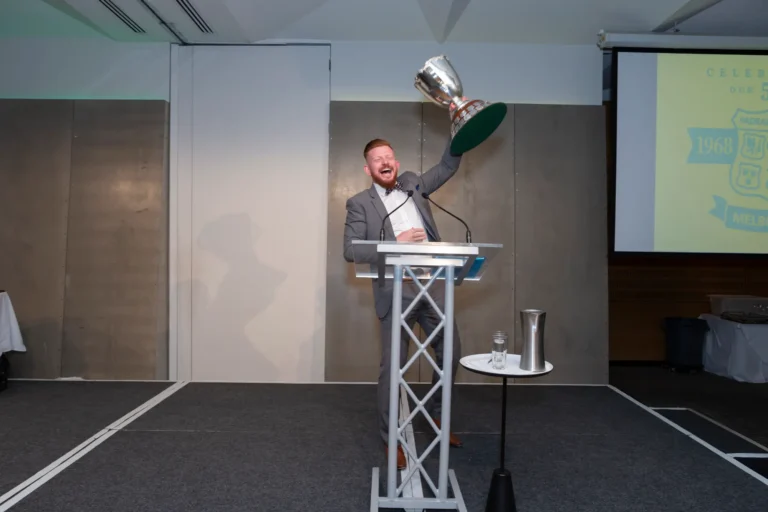 A man stands on a stage at the Pearses 50th Anniversary Awards Night at Zinc, Fed Square, celebrating a victory. Dressed in a gray suit and white shirt, he smiles broadly and raises a large silver trophy with a green base in his right hand while resting his left on a modern metal podium. Behind him, a projection screen displays "CELEBRATING OUR 50 YEARS 1968 MELBOURNE." The stage features gray and white paneled walls, a small table with a glass of water, and a dark gray carpeted platform. The moment captures energetic celebration and triumph.