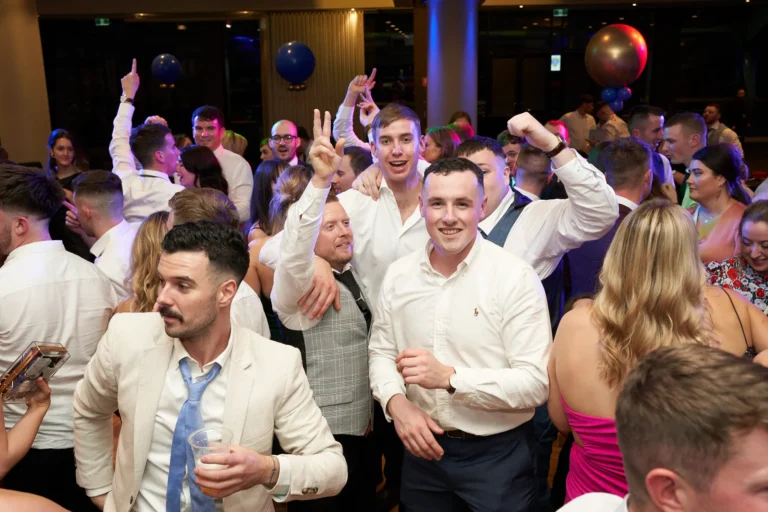 This image captures the joyous energy of St. Kevin’s 65th Anniversary Ball, with a group of men in formal wear leading the celebration. In the foreground, a man in a light suit jacket and blue tie holds a drink, while behind him, another man in a white shirt smiles at the camera, joined by a friend flashing a peace sign. Further back, another guest raises his arm in excitement. The dance floor is filled with well-dressed attendees dancing and mingling, with large balloons, including blue and red-gold ones, enhancing the festive and lively atmosphere of the anniversary celebration.