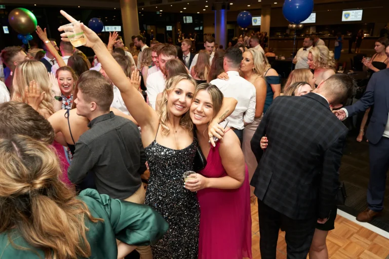 This image captures the vibrant atmosphere of St. Kevin’s 65th Anniversary Ball, with two women smiling brightly in the foreground. One wears a shimmering black sequined gown and raises a drink, her arm around a friend in a striking fuchsia dress holding a glass. Behind them, a lively crowd of men in suits and women in elegant gowns fill the dance floor, dancing and socializing. Their joyful expressions reflect the high-spirited energy of the evening. Large blue and green balloons and warm ambient lighting enhance the festive setting, creating a vivid scene of shared celebration and anniversary revelry.