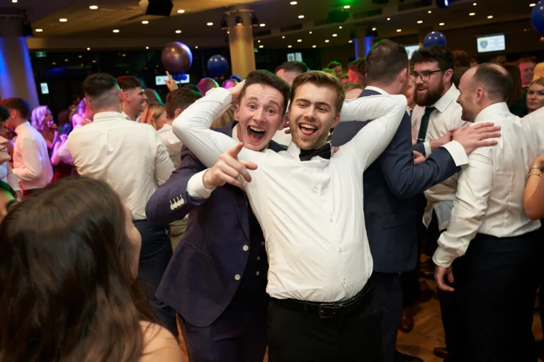 This image captures a joyful moment on the dance floor at St. Kevin’s 65th Anniversary Ball in Marvel Stadium. In the foreground, two men in formal attire celebrate enthusiastically—one in a dark suit laughing and pointing at the camera, while the other in a white shirt and black bow tie smiles broadly with arms raised behind his head. Behind them, the dance floor is crowded with elegantly dressed guests mingling, dancing, and enjoying the lively atmosphere. Balloons and ambient lighting enhance the celebratory mood, conveying a sense of fun, camaraderie, and shared joy during the milestone anniversary event.