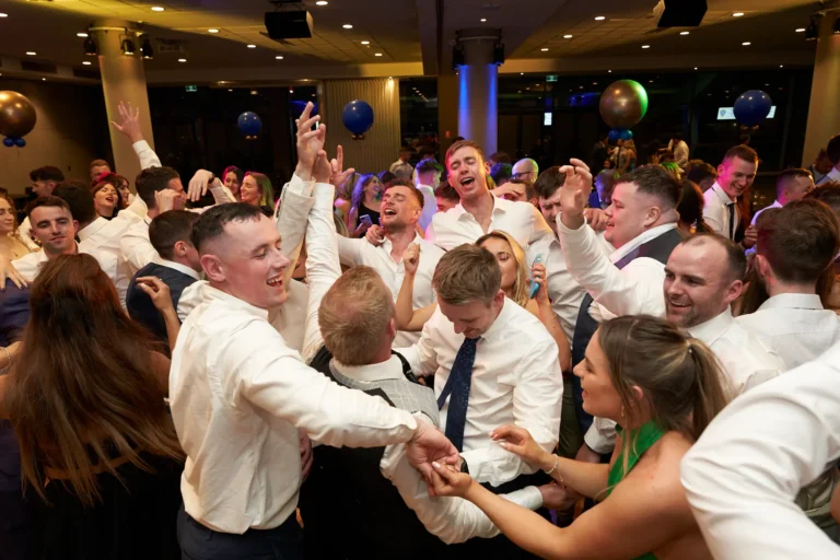 This image captures the exhilarating energy of the dance floor at St. Kevin’s 65th Anniversary Ball, where a lively crowd of young adults celebrates with unrestrained joy. Men in white shirts and ties and women in elegant dresses raise their arms, laugh, and cheer in rhythm with the music. A central group of men displays animated expressions, while women nearby in green and dark gowns join in the revelry. The background reveals a packed ballroom with festive lighting and decorative blue and gold balloons, enhancing the vibrant, communal spirit of this milestone celebration.