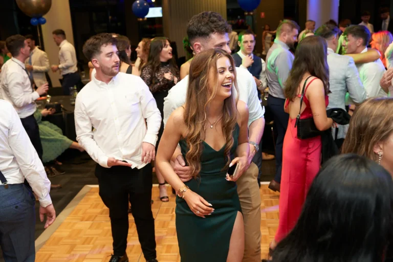 This image captures a lively moment on the dance floor during St. Kevin’s 65th Anniversary Ball at Marvel Stadium. A young woman in a dark green off-the-shoulder gown with a high slit laughs joyfully as a man in a light dress shirt embraces her from behind, both smiling. Surrounding them, other elegantly dressed guests mingle, dance, and enjoy the celebration. Balloons can be seen above, adding to the festive decor, while the patterned dance floor reflects the event lighting. The overall atmosphere conveys joy, camaraderie, and the vibrant spirit of the milestone anniversary celebration.