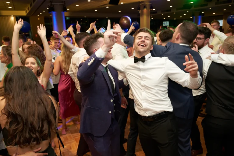 This image captures the joyous spirit of St. Kevin’s 65th Anniversary Ball at Marvel Stadium, focusing on a lively dance floor scene. In the foreground, two men in formal attire lead the celebration—one in a dark suit raising his hands enthusiastically, while the other, in a white dress shirt and bow tie, laughs with his head tilted back and arms lifted in joy. Surrounding them, a crowd of elegantly dressed guests dances, mingles, and enjoys the music. Festive decorations, balloons, and ambient lighting enhance the celebratory mood, reflecting the energy, camaraderie, and excitement of this milestone anniversary event.