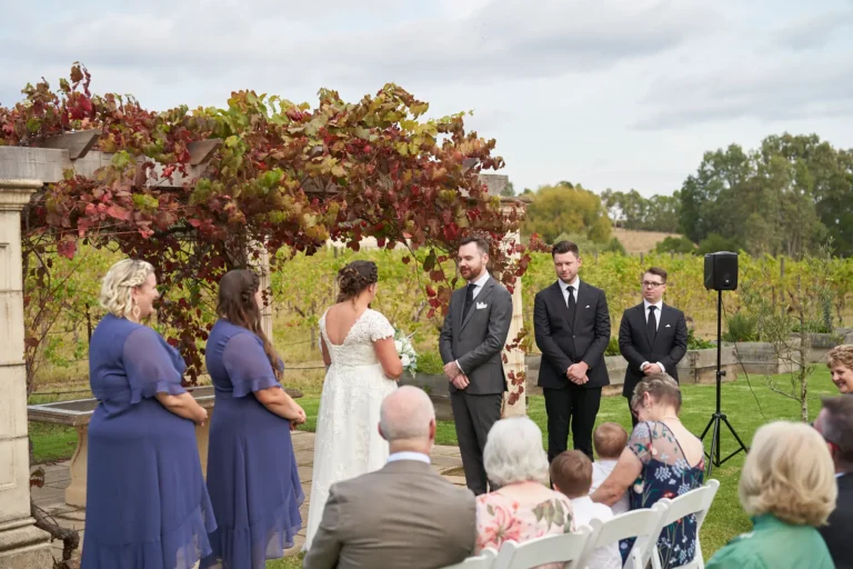 erin jesse bride groom standing ceremony proceeding sandstone gazebo fergusson winery yarra valley Bride and groom standing together at the Sandstone Gazebo during their outdoor wedding ceremony at Fergusson Winery in the Yarra Valley, facing each other with gentle smiles. The female officiant stands between them guiding the proceedings, while bridesmaids in blue dresses and groomsmen in dark suits stand nearby. Guests are seated on white chairs on the green lawn, attentively watching. The rustic stone gazebo is draped in lush red and green autumn grapevines, with rows of vineyards and rolling hills extending into the distance, creating a serene, intimate, and elegantly rustic atmosphere for this meaningful moment before the ring exchange.