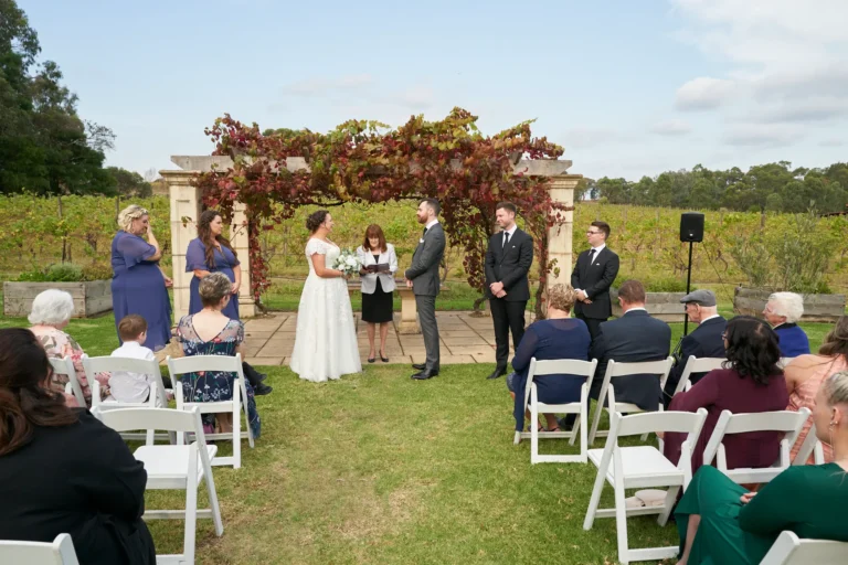 erin jesse bride groom standing ceremony sandstone gazebo fergusson winery yarra valley Bride and groom standing face-to-face at their wedding ceremony beneath the Sandstone Gazebo at Fergusson Winery in the Yarra Valley, looking at each other with gentle smiles. The officiant stands between them guiding the proceedings, while bridesmaids in blue dresses and groomsmen in dark suits stand nearby and guests watch from white chairs on the green lawn. The rustic stone gazebo is draped in lush red and green autumn grapevines, with rows of vineyards and rolling hills in the background, creating a serene, intimate, and elegantly rustic atmosphere for this moment before the exchange of rings.