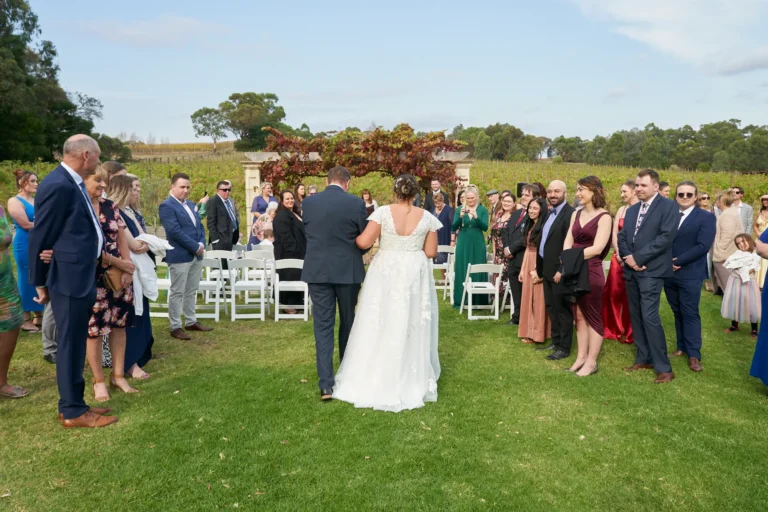 erin jesse bride walking aisle sandstone gazebo fergusson winery yarra valley Bride walking down the aisle on the arm of a man in a blue suit at Fergusson Winery in the Yarra Valley, approaching the Sandstone Gazebo for the outdoor ceremony. Guests stand along the green lawn, smiling and watching as the bride progresses. The rustic stone gazebo is draped in red and green autumn grapevines, with rows of vineyards and rolling hills in the background. The moment captures joyful anticipation, serene elegance, and the start of the wedding ceremony amid the picturesque vineyard setting.