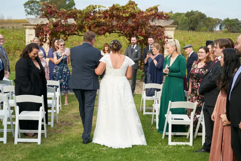 erin jesse bride walking aisle with father sandstone gazebo fergusson winery Bride walking down the aisle with her father at Fergusson Winery in the Yarra Valley, approaching the Sandstone Gazebo for the outdoor wedding ceremony. The bride wears a flowing white gown while her father escorts her on the green lawn lined with white guest chairs. Guests stand along the aisle, smiling and watching the processional. The rustic stone gazebo is draped in vibrant red and green autumn grapevines, with rows of vineyards and rolling hills visible in the distance, creating a heartfelt, joyful, and picturesque moment as the ceremony begins.