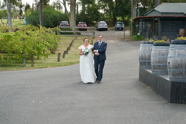 erin jesse bride walking with father driveway fergusson winery Bride walking arm-in-arm with her father down a paved driveway at Fergusson Winery in the Yarra Valley, en route from the guesthouse to the main wedding venue. The bride wears a white lace gown and holds a white and green bouquet, while her father is in a dark navy suit. Parked cars, vineyards, and the winery building with wine barrels and floral arrangements are visible, creating a serene, intimate, and rustic pre-ceremony moment filled with anticipation and joy.