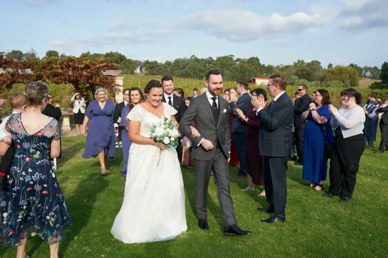 erin jesse couple joyful walking aisle post ceremony fergusson winery yarra valley Newly married couple walking arm-in-arm down the aisle immediately after their outdoor ceremony at Fergusson Winery in the Yarra Valley. Guests stand along the green lawn, smiling and clapping as the couple shares joyful expressions while walking away from the Sandstone Gazebo draped in lush red and green autumn grapevines. Rows of vineyards and rolling hills stretch into the distance under a partly cloudy blue sky, creating a celebratory, picturesque, and elegantly rustic moment that captures the happiness and excitement of the wedding day.