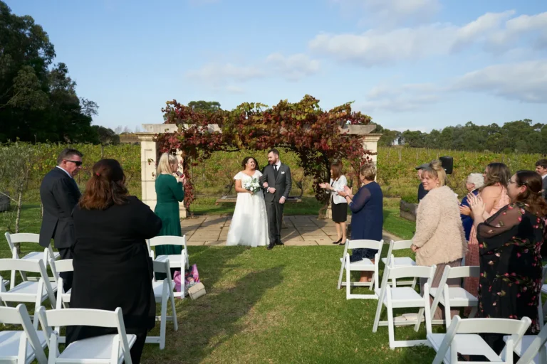 erin jesse couple walking aisle post ceremony fergusson winery yarra valley Newly married couple walking hand-in-hand down the aisle after their outdoor ceremony at Fergusson Winery in the Yarra Valley. Guests stand along the green lawn, clapping and smiling as the couple walks away from the Sandstone Gazebo draped in lush red and green autumn grapevines. Rows of vineyards and rolling hills stretch into the distance under a partly cloudy blue sky, creating a joyful, celebratory, and picturesque moment that blends rustic elegance with the serene beauty of the winery setting.