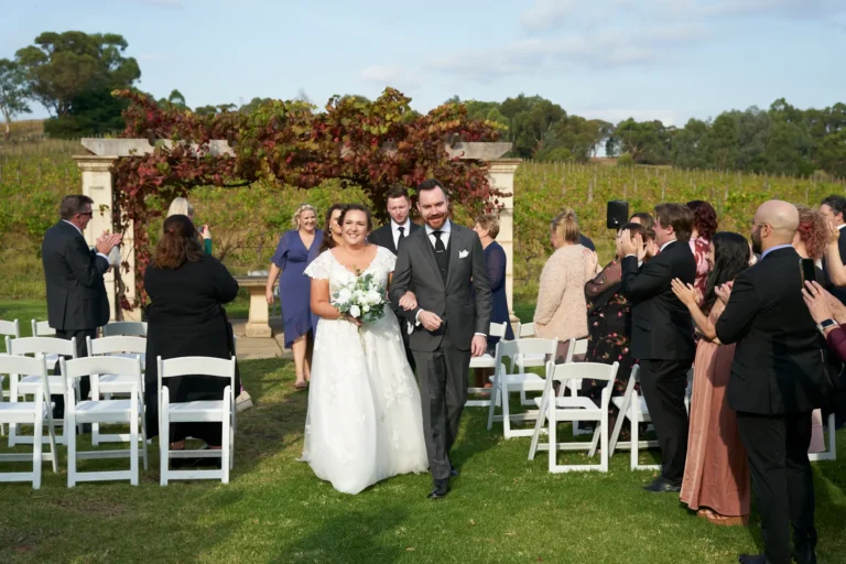 erin jesse couple walking aisle wedding party sandstone gazebo fergusson winery yarra valley Newly married couple walking arm-in-arm down the aisle at Fergusson Winery in the Yarra Valley, just after their outdoor ceremony at the Sandstone Gazebo. Guests line the green lawn, smiling and clapping, while members of the wedding party in blue dresses and dark suits walk just behind them, sharing in the joy. The gazebo, with light-colored stone pillars and a roof draped in red and green autumn grapevines, frames the couple. Rows of vineyards and rolling hills stretch into the distance under a partly cloudy blue sky, creating a festive, picturesque, and elegantly rustic atmosphere celebrating the couple’s first moments as husband and wife.
