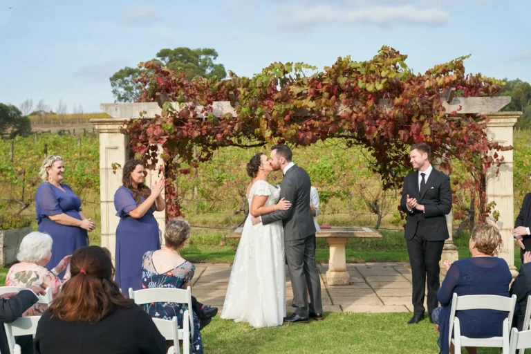 erin jesse first kiss sandstone gazebo fergusson winery yarra valley Bride and groom sharing their first kiss as a married couple beneath the Sandstone Gazebo at Fergusson Winery in the Yarra Valley. The groom’s left arm wraps around the bride’s waist while her left arm rests around his neck, capturing a romantic and intimate moment. Bridesmaids, groomsmen, and seated guests clap and smile in celebration. The rustic stone gazebo is draped in vibrant red and green autumn grapevines, with rows of vineyards and rolling hills in the background, creating a joyful, picturesque, and elegantly rustic atmosphere that marks the culmination of their outdoor wedding ceremony.