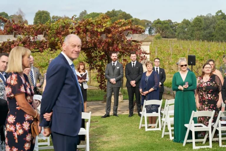 erin jesse groom and guests waiting ceremony fergusson winery Groom and groomsmen standing at the front of the aisle, awaiting the bride's arrival at Fergusson Winery in the Yarra Valley. Guests, dressed formally, are seated or standing along the grassy aisle, all focused on the entrance. A rustic pergola draped in red and green grapevines frames the ceremony area, with vineyards and rolling hills visible in the distance, creating a calm, formal, and anticipatory atmosphere for the outdoor wedding.