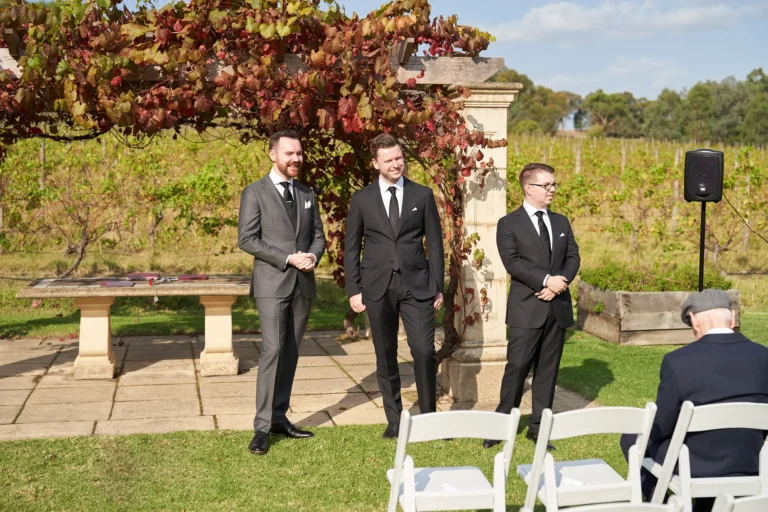 erin jesse groom groomsmen awaiting ceremony fergusson winery yarra valley Groom and groomsmen standing in a line, smiling and poised as they await the start of the outdoor ceremony at Fergusson Winery in the Yarra Valley. They stand on a paved stone area next to a green lawn with white guest chairs, in front of a rustic pergola draped with red and green autumn grapevines. Rows of vineyards and rolling hills stretch into the distance. Their relaxed, anticipatory expressions convey calm excitement, set within a serene, elegant, and picturesque vineyard backdrop that blends rustic charm with wedding-day formality.