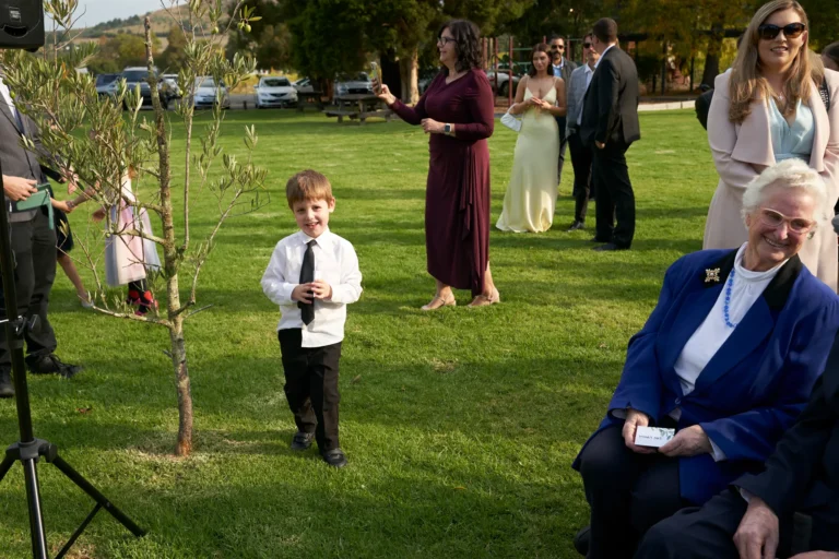 erin jesse guests gathering lawn fergusson winery yarra valley Guests mingling and waiting on the green lawn at Fergusson Winery in the Yarra Valley before the outdoor wedding ceremony begins. A young boy in a white shirt and black tie smiles at the camera in the foreground, while women in maroon, blue, yellow, and pink attire chat and observe the surroundings. The lush lawn is framed by trees, rolling hills, and a parking area in the background. The bright natural light and clear sky create a calm, social, and anticipatory atmosphere as guests gather in a relaxed and picturesque winery setting before the ceremony starts.