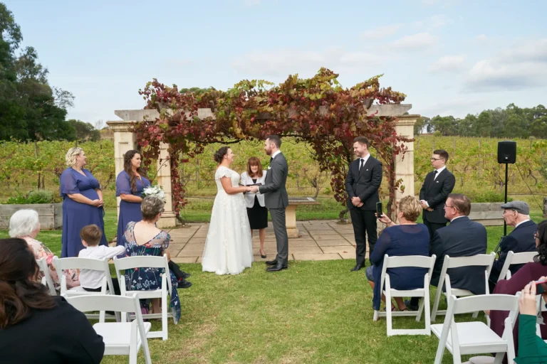erin jesse holding hands sandstone gazebo fergusson winery yarra valley Bride and groom holding hands each other hand, on the bride’s left finger beneath the Sandstone Gazebo at Fergusson Winery in the Yarra Valley. They smile gently at each other while the officiant stands between them guiding the ceremony. Bridesmaids in blue dresses and groomsmen in dark suits stand nearby as seated guests watch. The rustic stone gazebo is draped in lush red and green autumn grapevines, with rows of vineyards and rolling hills extending into the distance, creating an intimate, joyful, and elegantly rustic atmosphere for this meaningful outdoor wedding moment.