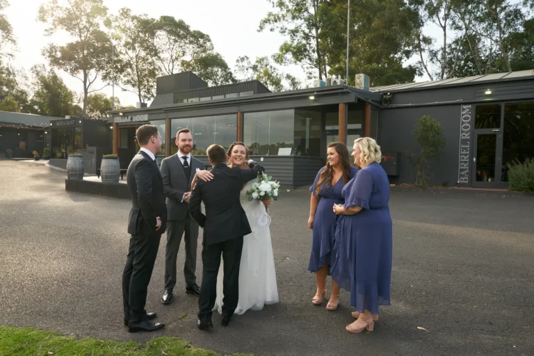 erin jesse post ceremony congratulations fergusson winery yarra valley Bride and groom sharing a warm embrace with a guest in a dark gray suit immediately after their ceremony at Fergusson Winery in the Yarra Valley. The groom faces the camera while the bride has her back to it, surrounded by smiling bridesmaids in navy blue dresses and groomsmen in suits. They stand on a paved area in front of the winery’s main building, with dark exterior walls, large glass windows, and visible “CELLAR DOOR” and “BARREL ROOM” signage. Natural sunlight illuminates the scene, creating a joyful, relaxed, and celebratory atmosphere that highlights intimate post-ceremony interactions.