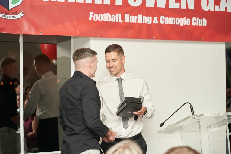 garryowens 65th anniversary ball award handshake During Garryowen’s 65th Anniversary Ball and awards night, a man receives two black presentation box awards from a speaker, exchanging a warm handshake. The winner smiles as he accepts the recognition, while the presenter looks on with a friendly, professional demeanor. The indoor venue features a polished floor, a clear acrylic podium, and a prominent red banner reading “GARRYOWEN G.A.C Football, Hurling & Camogie Club.” Other guests, seated and standing at formally set tables, observe the ceremony. Red balloons in the background add a festive touch, capturing a formal, joyful, and celebratory moment of recognition during the awards gala.