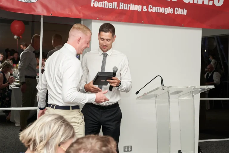 garryowens 65th anniversary ball award presentation men During Garryowen’s 65th Anniversary Ball and awards night, a man receives a black presentation box award from a speaker at a clear acrylic podium. The award recipient smiles broadly, looking down at the box, while the presenter observes the exchange. Guests in formal attire are seated at tables in the background, watching the ceremony. The polished indoor venue features a prominent red banner displaying “GARRYOWEN Football, Hurling & Camogie Club,” white walls, and a festive red balloon. This image captures a joyful, formal moment of recognition, reflecting the celebratory, professional, and sophisticated atmosphere of the milestone awards gala.