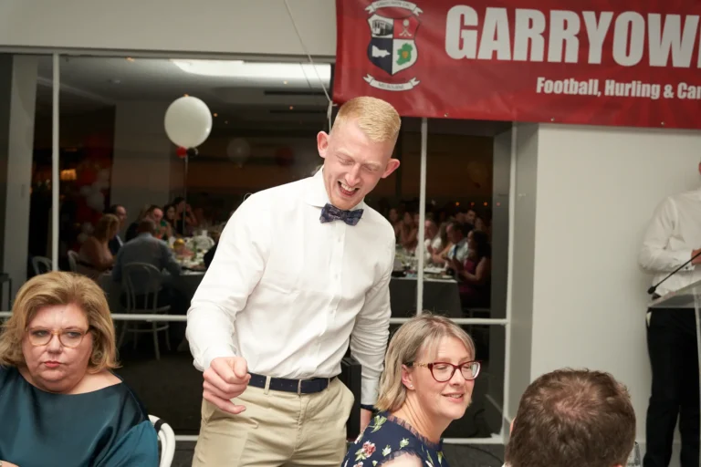 garryowens 65th anniversary ball award recipient excited A young man in a white shirt, beige trousers, and dark bow tie reacts with wide, joyful laughter as he approaches the stage during Garryowen’s 65th Anniversary Ball and awards night. Seated guests at nearby tables, including women in teal and floral dresses, smile and watch his exuberant reaction. The bright indoor venue features a large red banner with “GARRYOWEN Football, Hurling & Camogie Club,” round dining tables, and a floating white balloon. The scene captures a lively, festive, and social atmosphere, conveying excitement, celebration, and shared happiness during the milestone awards ceremony.