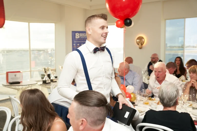 garryowens 65th anniversary ball award recipient smiling A young man dressed in a white shirt, blue suspenders, and bow tie smiles broadly as he holds a black presentation box during Garryowen’s 65th Anniversary Ball. Standing near a dining table, he appears to be returning to his seat after receiving an award. Guests in formal attire are seated nearby, observing and engaging in conversation. The bright, modern venue features polished hardwood floors, panoramic waterfront views through large windows, and festive red and black balloons. Silver trophies and elegantly set tables with white linens complete the sophisticated setting, capturing the pride, joy, and celebratory energy of the awards ceremony.