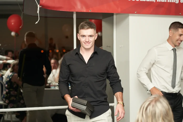 garryowens 65th anniversary ball award winner posing A young man proudly poses for the camera while walking back to his table at Garryowen’s 65th Anniversary Ball and awards night. Dressed in a black long-sleeved shirt and light plaid trousers, he holds two black presentation box awards in his left hand, smiling broadly. The modern venue features a polished hardwood floor, a red banner with the club crest, and festive red balloons. Seated guests at white-clothed tables watch the proceedings. This image captures a joyful, celebratory, and sophisticated moment of recognition, pride, and camaraderie during the formal awards ceremony.