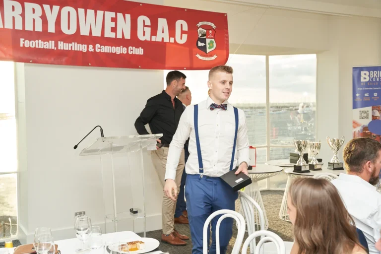 garryowens 65th anniversary ball award winner walking An award winner walks back to his seat during Garryowen’s 65th Anniversary Ball, smiling warmly while holding a small black presentation box. Dressed in a white shirt, blue trousers, bow tie, and suspenders, he passes seated and standing guests observing the proceedings. Behind him, a man stands near a clear acrylic podium, with silver trophies displayed on a contemporary white table and a large red “GARRYOWEN G.A.C Football, Hurling & Camogie Club” banner overhead. Large windows illuminate the modern venue, with polished hardwood floors and elegantly set tables, capturing a joyful, celebratory, and professional awards night atmosphere.
