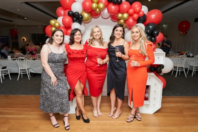 garryowens 65th anniversary ball awards night five women group posing Five elegantly dressed women pose side-by-side in front of a vibrant balloon arch and large illuminated "65" numbers at Garryowen’s 65th Anniversary Ball. Smiling warmly at the camera, the women hold glasses of wine or champagne, suggesting a joyful toast. The polished indoor venue, with set tables and a distant bar area, adds context to the lively gala. Their confident, relaxed poses and the festive decorations capture a moment of camaraderie, celebration, and sophistication, highlighting the excitement and warmth of this milestone awards night and anniversary event.