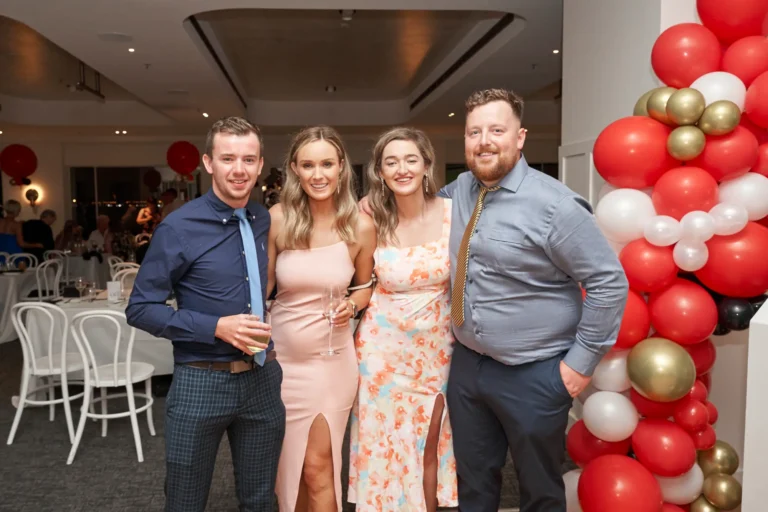 garryowens 65th anniversary ball awards night four friends posing Four friends, two men and two women, stand closely together smiling warmly at the camera during Garryowen’s 65th Anniversary Ball and awards night. Dressed in elegant formal attire, some holding glasses, they pose confidently yet relaxed. Behind them, a vibrant balloon arch in red, black, gold, and white and elegantly set dining tables highlight the festive indoor venue. The polished floor, soft lighting, and distant bar area add context to the lively gala. Their joyful expressions and the celebratory decor capture a sophisticated, happy, and memorable social moment at this milestone awards event.