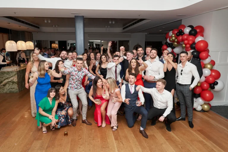 garryowens 65th anniversary ball awards night group photo A large group of around thirty elegantly dressed adults poses together during Garryowen’s 65th Anniversary Ball and awards night. Some kneel in front while others stand behind, ensuring everyone is visible. Guests smile, laugh, and interact warmly, many holding drinks, with one person raising an arm in celebration. The indoor venue features polished hardwood floors, soft ambient lighting, and a striking balloon arch in red, black, gold, and white. A bar area is visible in the mid-ground. The scene captures a lively, festive, and joyful atmosphere of camaraderie and shared celebration at this milestone event.