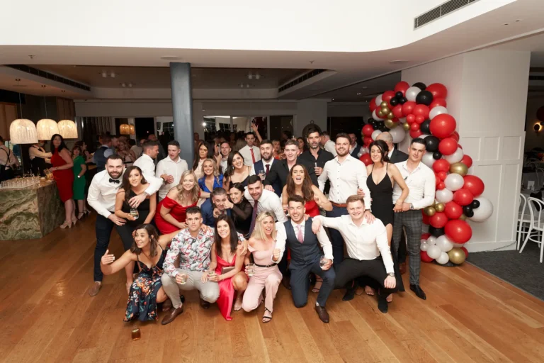 garryowens 65th anniversary ball awards night large group posing A large group of approximately thirty well-dressed adults poses together during Garryowen’s 65th Anniversary Ball and awards night. Arranged in multiple levels, some kneeling in front and others standing behind, they smile, laugh, and interact warmly. Many hold drinks, and one person raises an arm in celebration. The elegant indoor venue features polished hardwood floors, soft lighting, and a vibrant balloon arch in red, black, gold, and white, with a bar area visible in the mid-ground. The scene conveys a lively, festive, and memorable celebration of camaraderie and shared happiness.