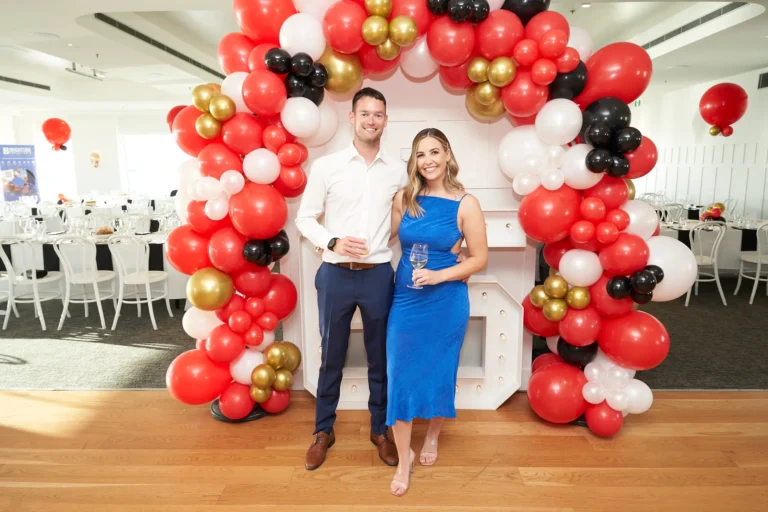 garryowens 65th anniversary ball couple arrival posing 65 backdrop A stylish couple poses during their arrival at Garryowen’s 65th Anniversary Ball, standing beside large illuminated “65” numbers framed by a festive balloon arch in red, black, gold, and white. The man, dressed in a white shirt and navy trousers, holds a drink while the woman in a vibrant blue slip dress smiles warmly with a wine glass in hand. Behind them, elegantly set dining tables with white linens and chairs hint at the formal celebration ahead. The polished hardwood floors and bright lighting enhance the chic, joyful, and celebratory atmosphere of the awards night.