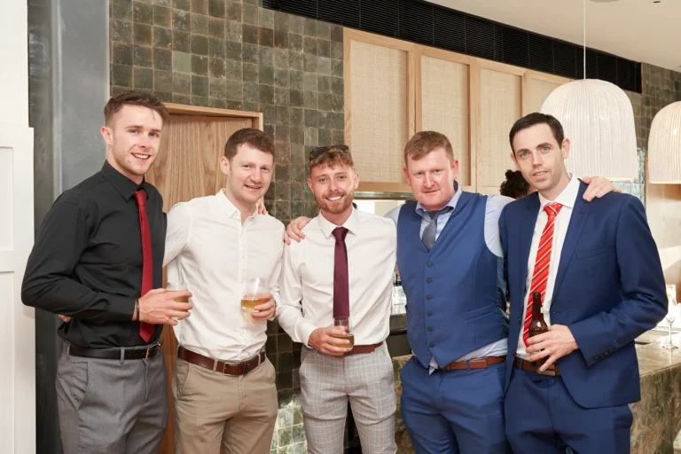 garryowens 65th anniversary ball five men arrival group photo Five men dressed in sharp suits and vests pose together during arrivals at Garryowen’s 65th Anniversary Ball, smiling warmly with drinks in hand. Standing close with arms around each other, they create a relaxed, friendly group moment that reflects the camaraderie of the evening. Behind them, the modern venue features textured dark tile, wood paneling with woven rattan, and a marble-topped bar beneath a large white spherical light. The scene captures the cheerful, social energy of the awards night as guests begin mingling and celebrating.