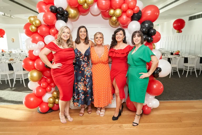 garryowens 65th anniversary ball five women arrival posing Five elegantly dressed women pose together during arrivals at Garryowen’s 65th Anniversary Ball, smiling brightly at the camera. One woman holds a small black clutch while the others stand confidently, framed by a festive balloon arch in red, white, black, and gold, alongside illuminated “65” numbers. Polished hardwood floors, well-set dining tables with white linens, and bright natural light from large windows create a stylish indoor waterfront venue. The scene captures a lively, chic, and celebratory atmosphere, reflecting the excitement, anticipation, and joyful social energy of guests at the milestone awards night.