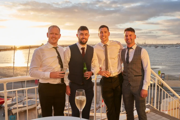 garryowens 65th anniversary ball four men balcony sunset Four well-dressed men pose together on the balcony of the Harbour Room during Garryowen’s 65th Anniversary Ball at sunset. Wearing formal attire, they smile broadly at the camera, with one man holding a wine glass, another a beer bottle, and a third a clear glass. The man on the right has his arm around his friend. Behind them, the golden glow of the setting sun illuminates Port Phillip Bay and the iconic St Kilda Pier, while red and white boats are docked below. The scene captures joyful camaraderie, sophistication, and a festive celebration during the gala event.