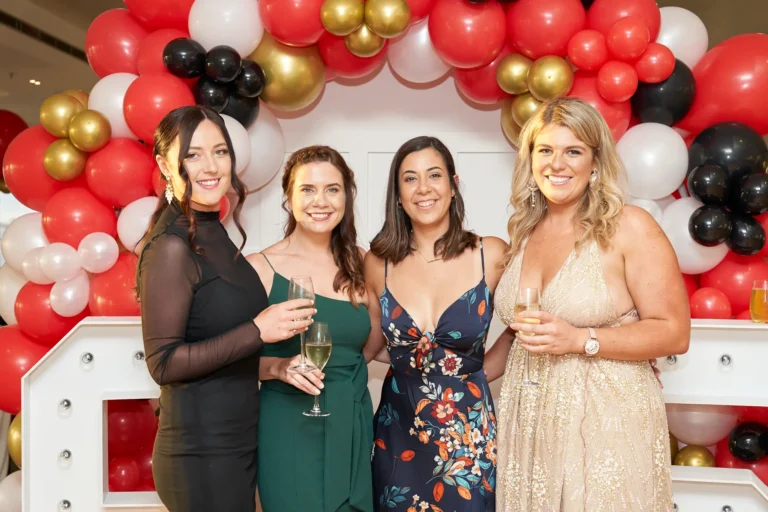 garryowens 65th anniversary ball four women balloon backdrop Four elegantly dressed women pose together at Garryowen’s 65th Anniversary Ball, each holding a glass of sparkling wine or champagne. They stand side-by-side, smiling brightly at the camera, wearing a mix of black, green, floral, and gold cocktail dresses. Behind them, a festive arch of red, white, gold, and black balloons frames the scene, with large illuminated “65” marquee numbers adding a celebratory touch. The neutral indoor wall enhances the vibrant decorations, creating a chic and joyful atmosphere. This image captures a glamorous, social, and lively moment of shared happiness during the milestone anniversary gala.