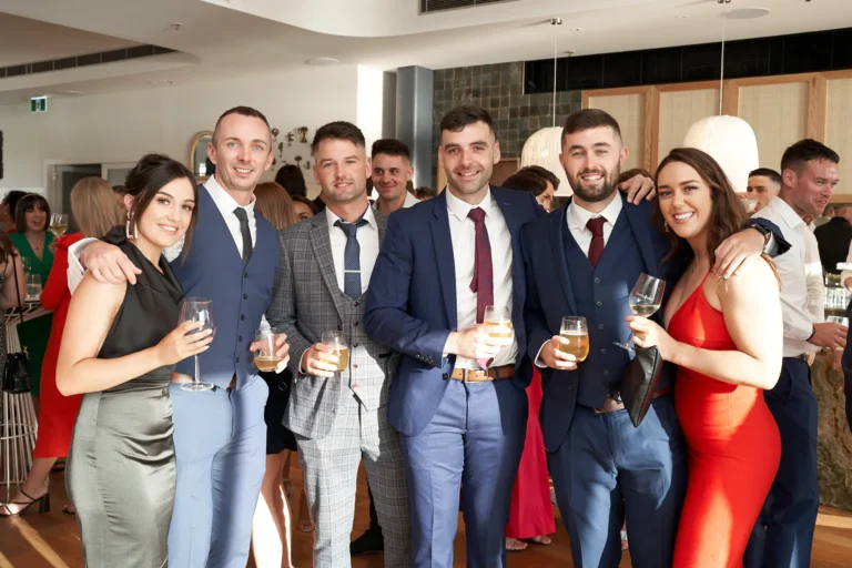 garryowens 65th anniversary ball group arrivals cheerful guest portrait A lively group of six elegantly dressed guests gather closely during arrivals at the Garryowen’s 65th Anniversary Ball. Smiling warmly at the camera, they hold wine and beer glasses, some with arms wrapped around each other in friendly celebration. Soft natural light from nearby windows blends with the venue’s warm indoor lighting, highlighting the polished wooden floors and modern architectural details. The atmosphere feels joyful, social, and full of anticipation as the evening’s formal festivities begin.