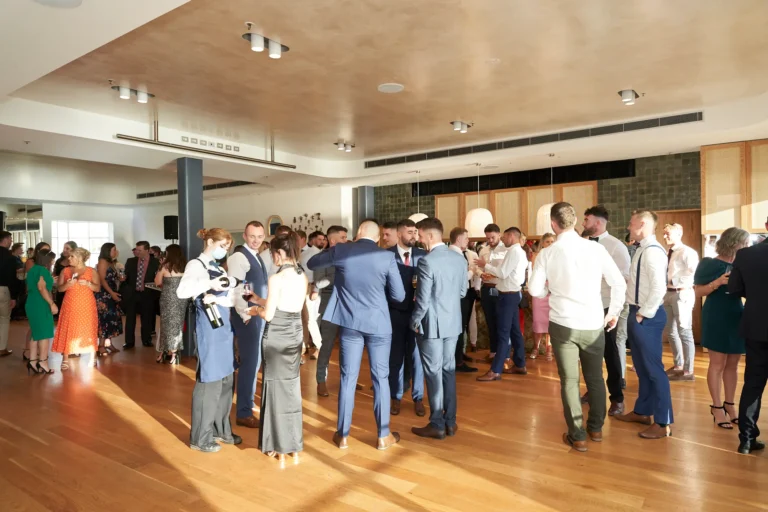 garryowens 65th anniversary ball guest arrivals mingling reception Guests mingle during the arrival hour of Garryowen’s 65th Anniversary Ball, dressed in elegant suits and cocktail attire. A central group of men huddles in conversation while others hold drinks, chat, and move through the bright, modern venue with polished hardwood floors, white and dark stone walls, and warm wooden paneling. Large windows flood the space with natural light, and a masked server offers refreshments in the background. The lively, bustling scene captures the excitement, social energy, and anticipation of the evening’s awards night, highlighting a well-attended, sophisticated, and joyful celebration.