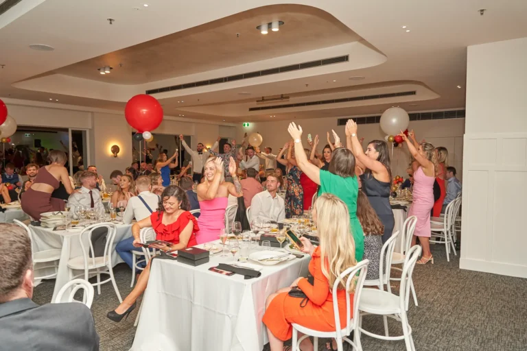 garryowens 65th anniversary ball guests celebrating award Guests react with exuberant excitement during Garryowen’s 65th Anniversary Ball and awards night. Many stand, cheer, and raise glasses, while seated guests smile and engage with the celebratory energy. A woman in a pink dress clasps her hands in delight, adding to the lively atmosphere. The elegant venue features curved ceilings, recessed lighting, dark carpeted floors, and festive red and white balloons. Tables are draped in white linens and surrounded by white chairs, with a distant bar area visible. This image captures a peak moment of joy, camaraderie, and collective celebration during the awards ceremony.