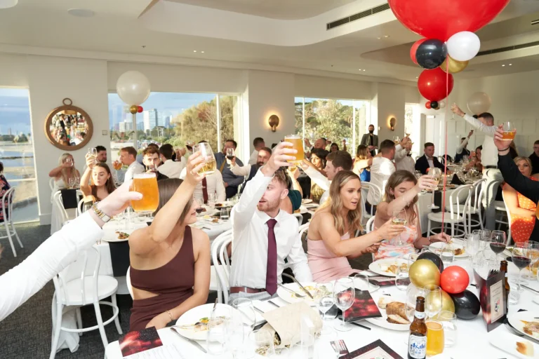 garryowens 65th anniversary ball guests toasting celebration Guests raise their glasses in a joyful toast during Garryowen’s 65th Anniversary Ball and awards night. Seated at elegantly set tables with white linens and multiple glasses, formally dressed attendees smile and engage with each other, celebrating a special moment. Floor-to-ceiling windows reveal panoramic views of a bay and outdoor deck, while large red, black, gold, and white balloons add festive flair. The polished floors, bright natural light, and elegant venue create a sophisticated backdrop. This scene captures camaraderie, excitement, and peak celebratory energy following an award presentation or special recognition during the milestone gala.