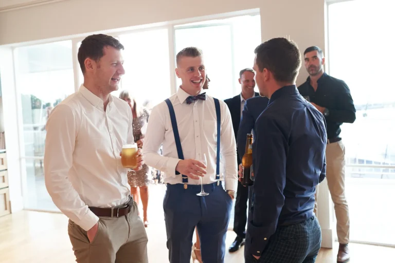 garryowens 65th anniversary ball men arrival socialising A group of men in formal and cocktail attire engage in lively conversation during guest arrivals at Garryowen’s 65th Anniversary Ball. Two men in the foreground smile at each other while others share laughter, holding drinks including glasses of beer and a brown bottle. The bright, modern venue features polished light hardwood floors, minimalist white walls, and large windows that fill the space with natural light, offering glimpses of the outdoor area. The scene captures a cheerful, sophisticated atmosphere of camaraderie, social energy, and anticipation as guests mingle before the awards night festivities begin.