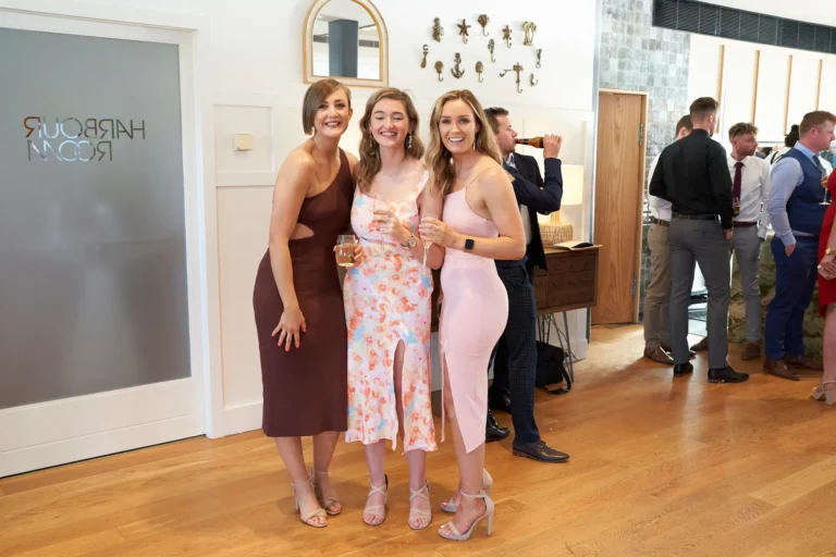 garryowens 65th anniversary ball three guests arrival posing Three women in elegant cocktail dresses pose together during guest arrivals at Garryowen’s 65th Anniversary Ball, smiling warmly as they hold their drinks and lean in for the photo. Behind them, a man takes a sip from a bottle while other formally dressed guests mingle and chat across the bright, stylish venue. Polished hardwood floors, mirrored doors, an arched wall mirror, and decorative artwork create an elegant backdrop, with warm lighting enhancing the lively reception atmosphere. The scene captures the excitement, social energy, and celebratory spirit welcoming guests to the awards night.