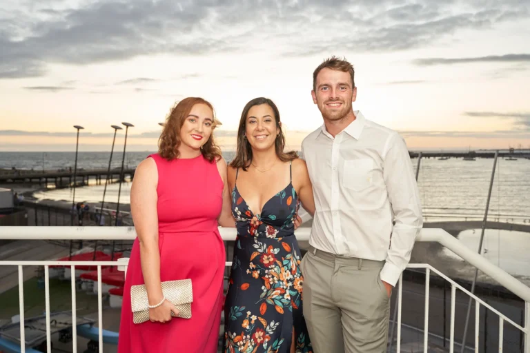 garryowens 65th anniversary ball three guests balcony sunset Three elegantly dressed adults—two women and one man—pose together on the balcony of the Harbour Room during Garryowen’s 65th Anniversary Ball at sunset. Smiling warmly at the camera, the woman in red holds a gold clutch, the woman in a floral dress links arms with the man in a white shirt and grey trousers. Behind them, Port Phillip Bay and St Kilda Pier glow under the golden sunset, with docked boats below. The image conveys joyful celebration, relaxed sophistication, and shared happiness during a vibrant, festive golden-hour moment.