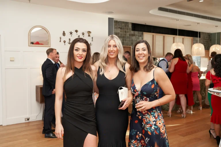 garryowens 65th anniversary ball three women arrival portrait Three elegantly dressed women pose together during guest arrivals at Garryowen’s 65th Anniversary Ball, smiling confidently at the camera. One wears a black cross-halter dress, the second a black v-neck dress, and the third a navy floral-print slip dress holding a wine glass. Behind them, formally dressed guests mingle near a bar in a modern, stylish venue with polished hardwood floors, white and dark stone accent walls, and elegant spherical pendant lighting. The warm lighting and vibrant interactions capture the joyful, sophisticated, and celebratory atmosphere of the evening’s formal awards night.