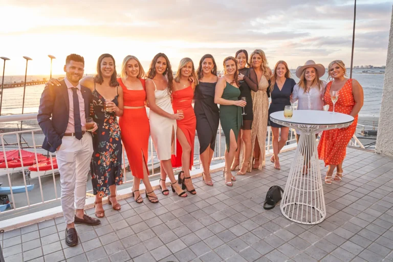 garryowens 65th anniversary ball twelve people balcony sunset A group of twelve elegantly dressed adults—eleven women and one man—pose together on the balcony of the Harbour Room during Garryowen’s 65th Anniversary Ball at sunset. Smiling broadly, some hold glasses of wine, beer, or cocktails, while the man has his arm around a woman. Behind them, Port Phillip Bay and St Kilda Pier are bathed in the golden light of the setting sun, with red and white boats docked below. The image captures joyful camaraderie, sophistication, and a warm, festive celebration during a stunning golden-hour moment.